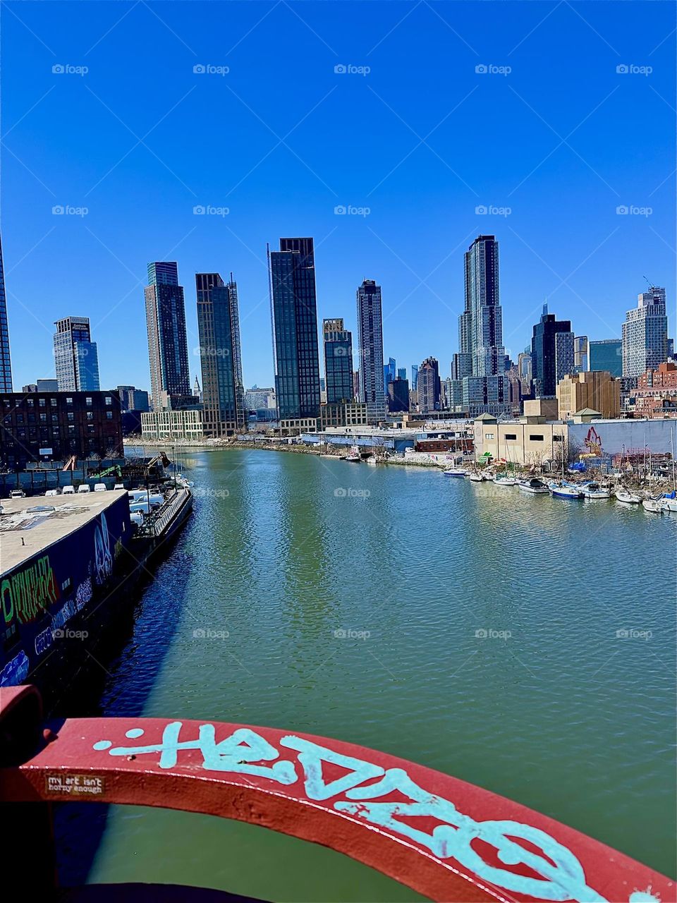 This is “Newtown Creek” seen from the “Pulaski Bridge” that connects LIC, Queens to “Greenpoint”, Brooklyn. The graffiti covered red metal railing wraps around one of the viewing platforms. 2024. Hypnotic Productions