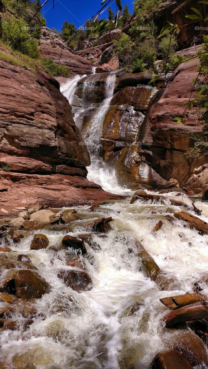 Red rocks glisten under sparkling waters of this beautiful waterfall on a blue sky summer day.