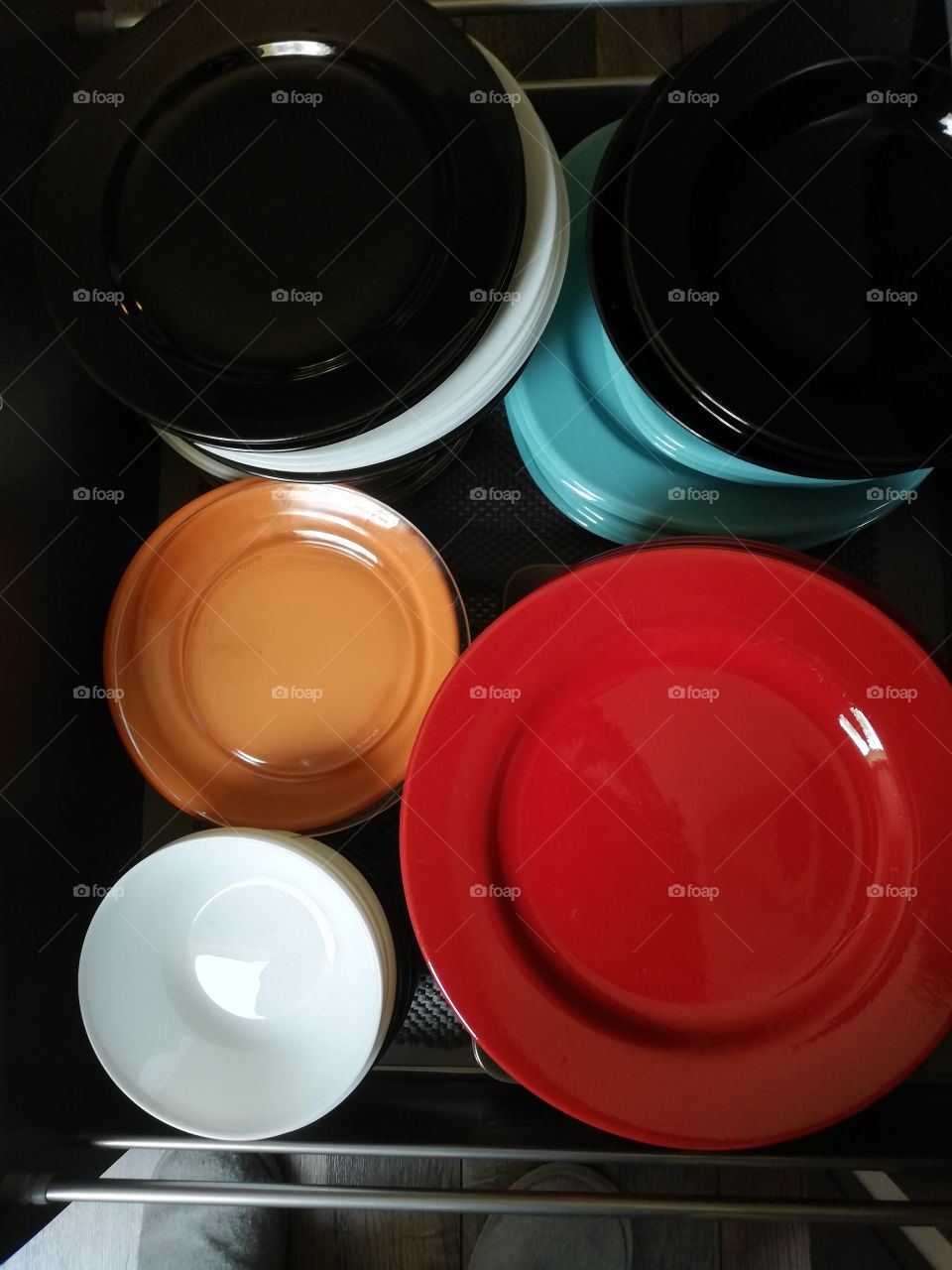 The plates in many colours and sizes are one on the other inside the cupboard, which walls are stuck with the parts of grey plastic and silver color metallic holders. The shoes are seen on the floor.