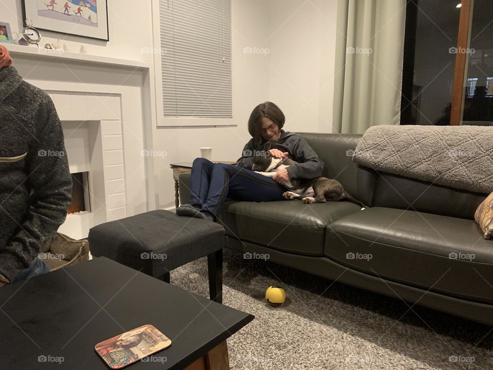 A Boston terrier and pug mix cuddling up with a woman on the couch 