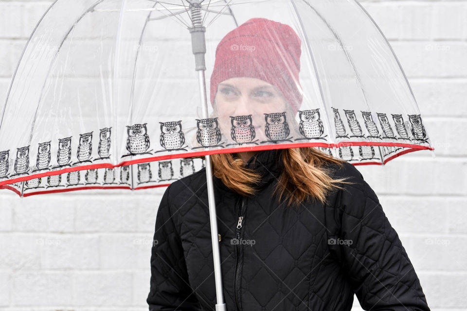 Young woman using a clear umbrella against a white brick wall