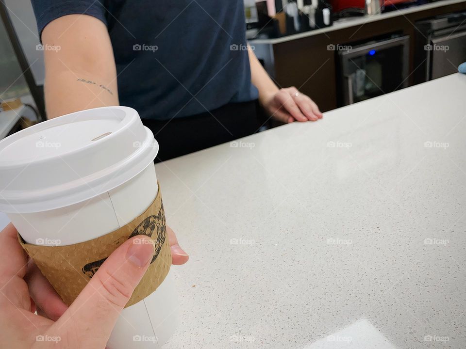 Handed a delightful coffee sweet latte in a disposable cup in the interior of a coffee shop by the coffee maker worker on the shiny counter.