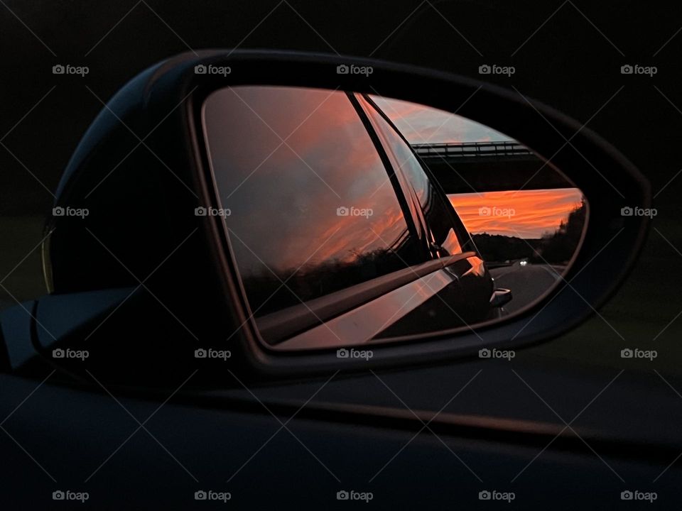 View of a bridge in the side mirror of a car at sunset. Another car‘s headlights can be seen in the back.