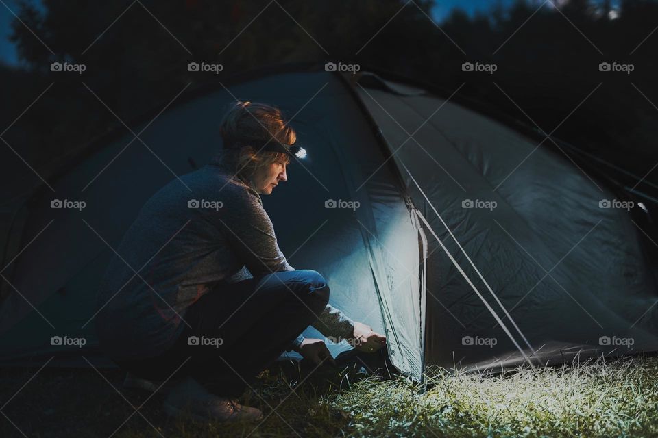 Summer vacation. Woman opening tent at camping in the evening using head lamp. Summer trip. Preparing campsite. Spending vacation outdoors close to nature