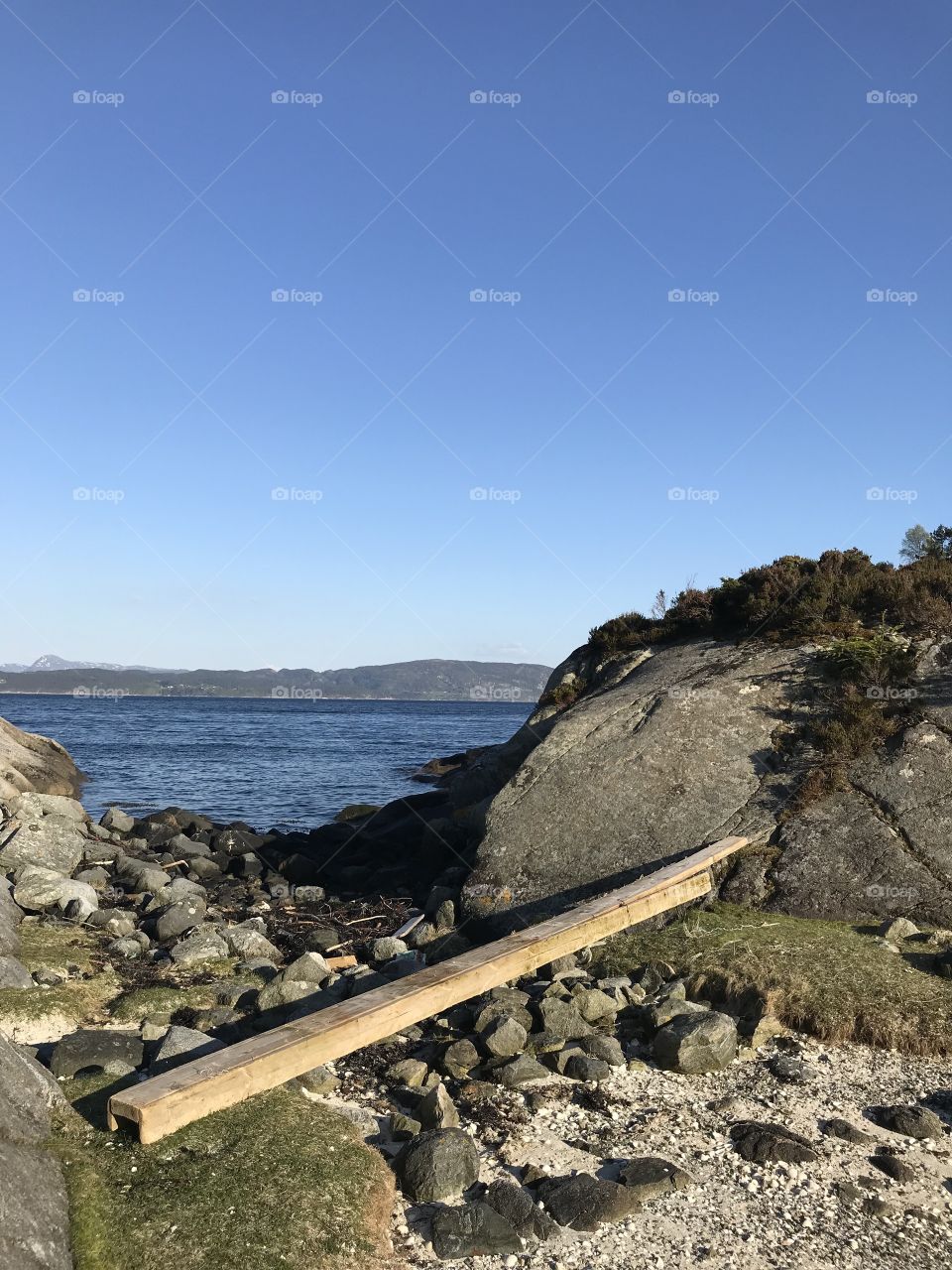 A plank laying on many rocks in Norway. Behind the plank it’s the ocean and landscape. 