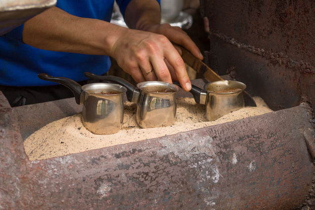 A man makes coffee on hot sand in a stainless turk