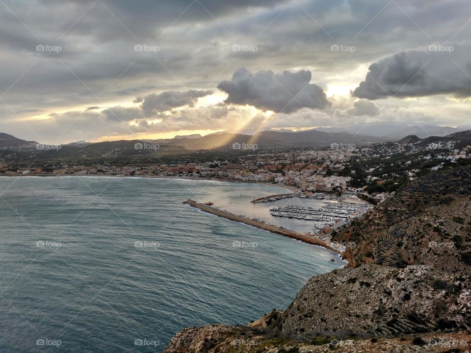 Landscape from the cliff, sea, city, green, and the sun's rays passing through some clouds and illuminating the mountains and the city of Javea in Spain.