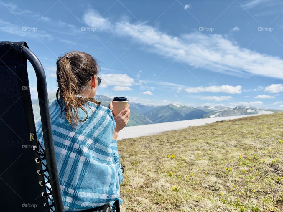 girl in a blanket with a cup of coffee sits in a chair in the mountains