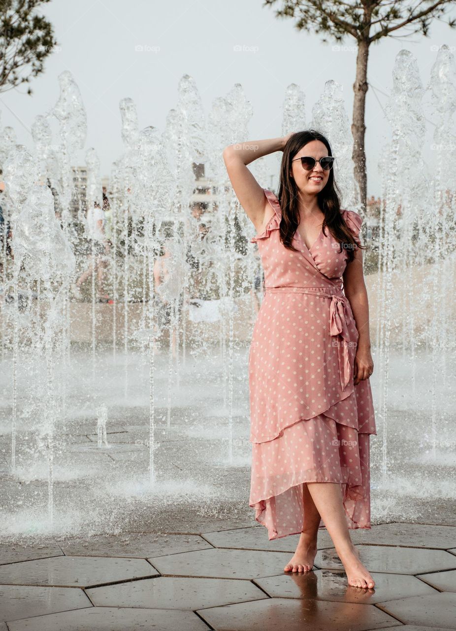 Full body portrait of young woman wearing a pink dress standing in front of a fountain in city on a hot summer day