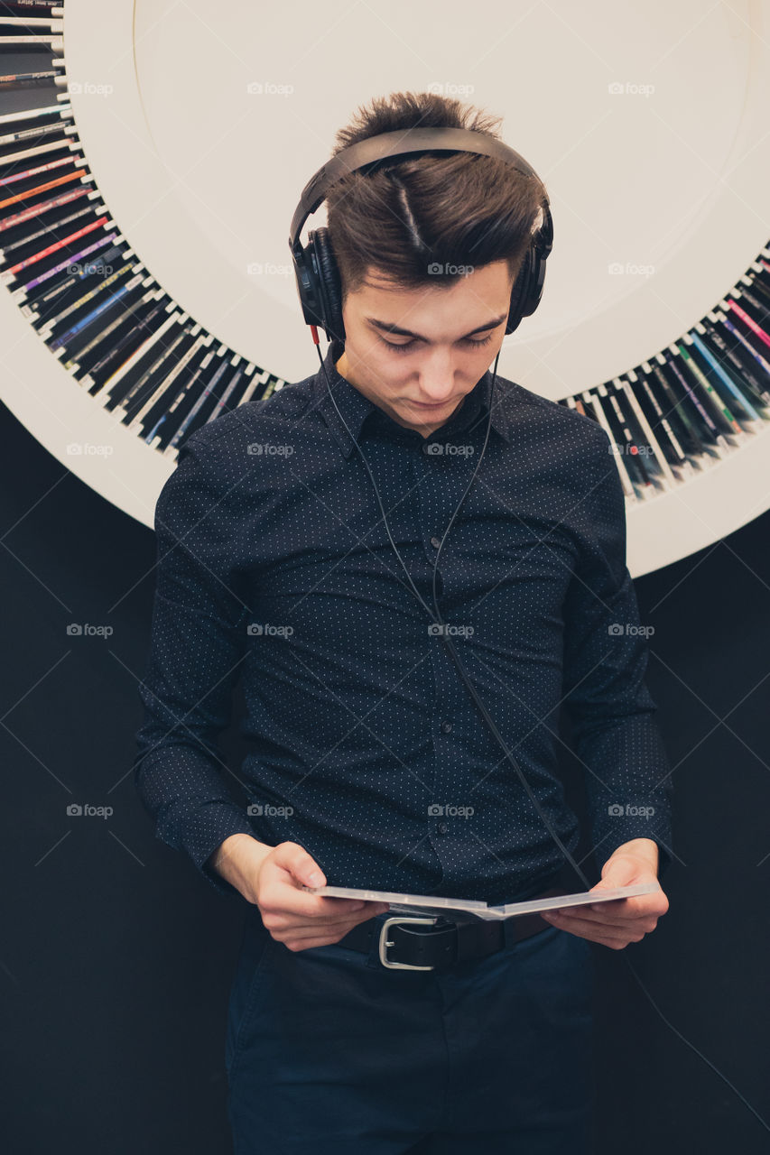 Young man listening to music through headphones standing next to bookshelf with recordings
