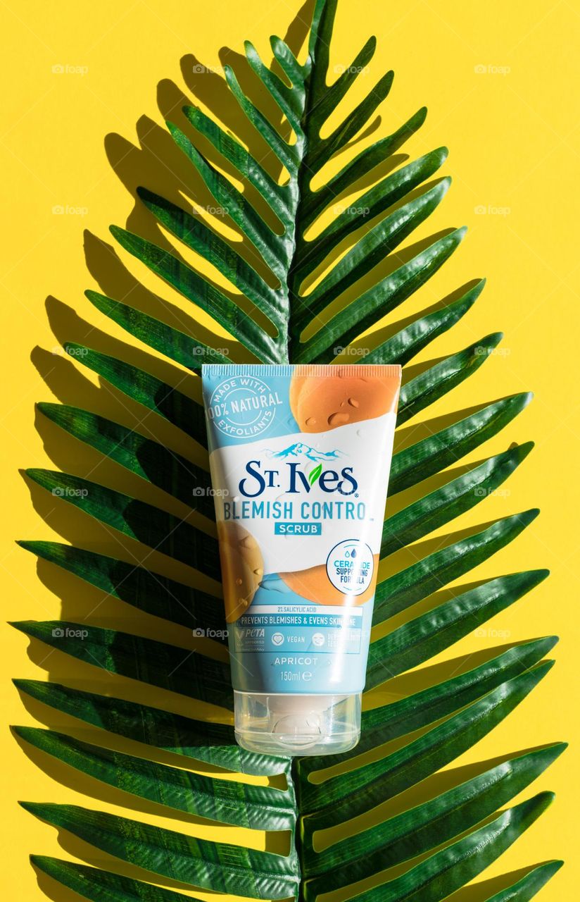 One tube natural apricot body scrub brand st. Ives lies on one palm branch with leaves in the center on a yellow background, flat lay close-up. The concept of female cosmetics, tie behind the body, beauty.