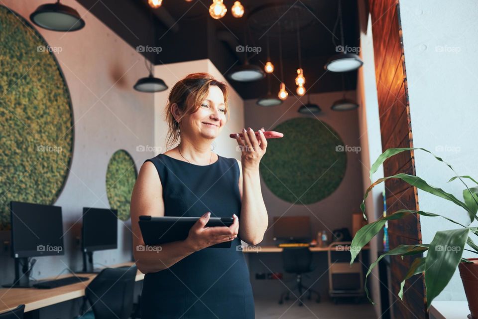 Businesswoman having business call working on tablet in office. Mature busy woman using touch pad computer standing by window in modern interior. Female manager focused on work holding digital device. Using technology