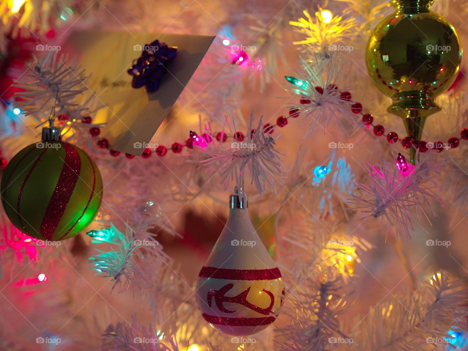 Christmas ornaments hang in a white tree which glows peacefully with multicolored lights. 
