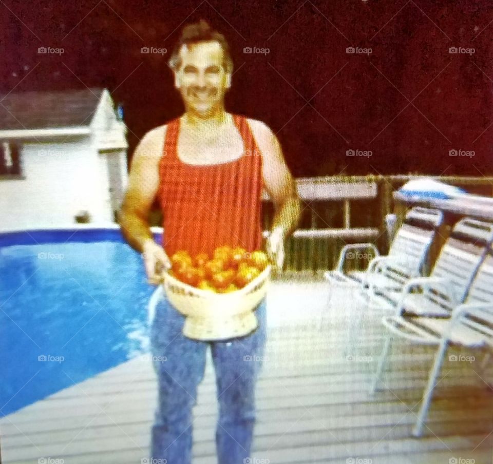 Man holding basket full of ripe tomatoes, pool & deck in photo.