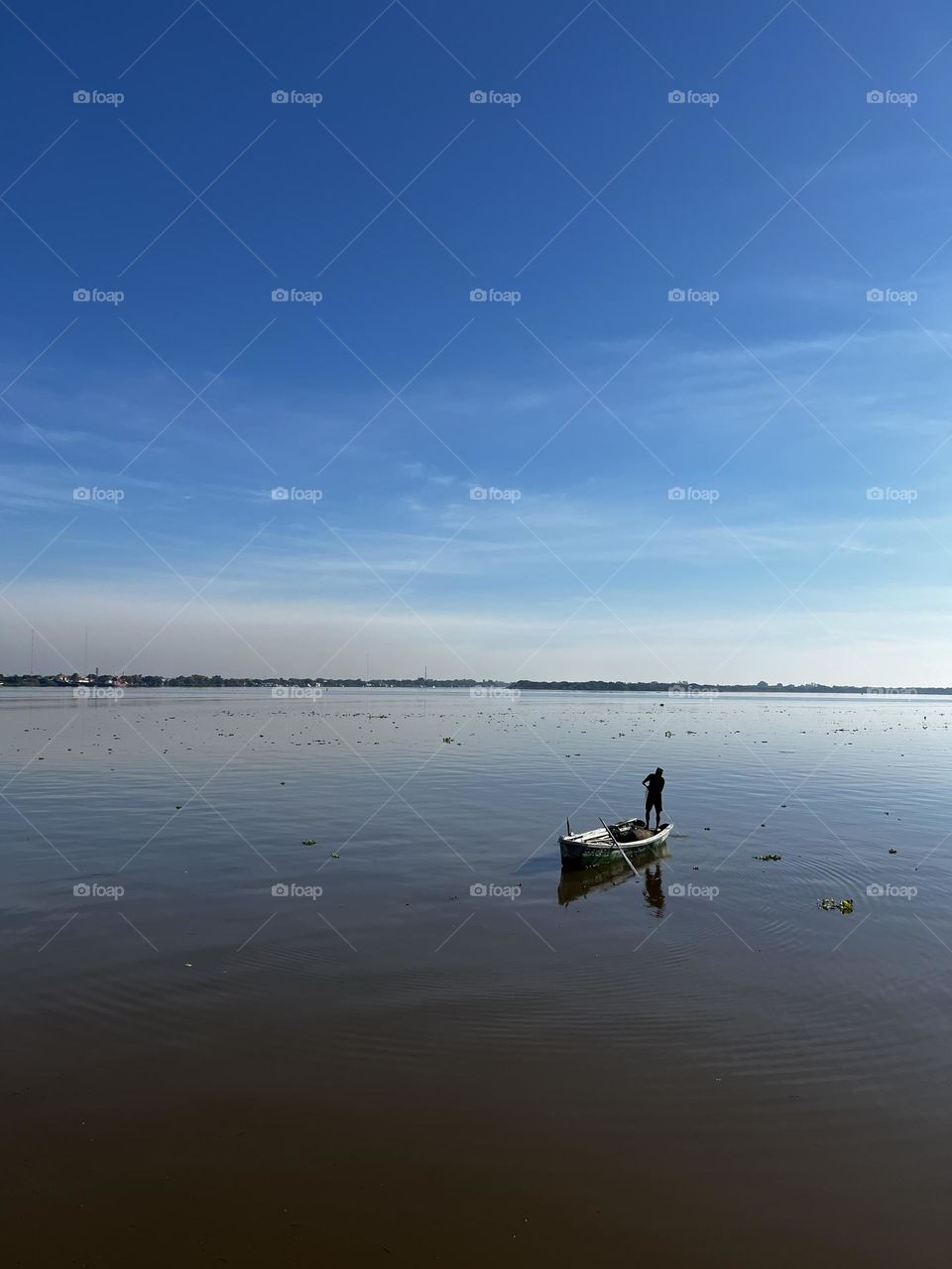 A lonely fisherman on a small vessel in the middle of a vast reflective pond under a clean and clear blue sky. The environment transmits tranquility and connection with nature.