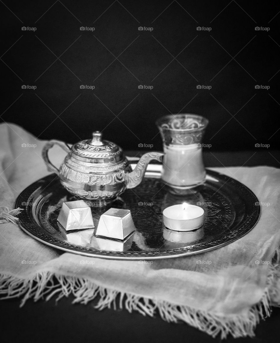 Silver ornamental tray with a small silver teapot and a glass with tea with milk, sweets and a candle on gray beautiful textile