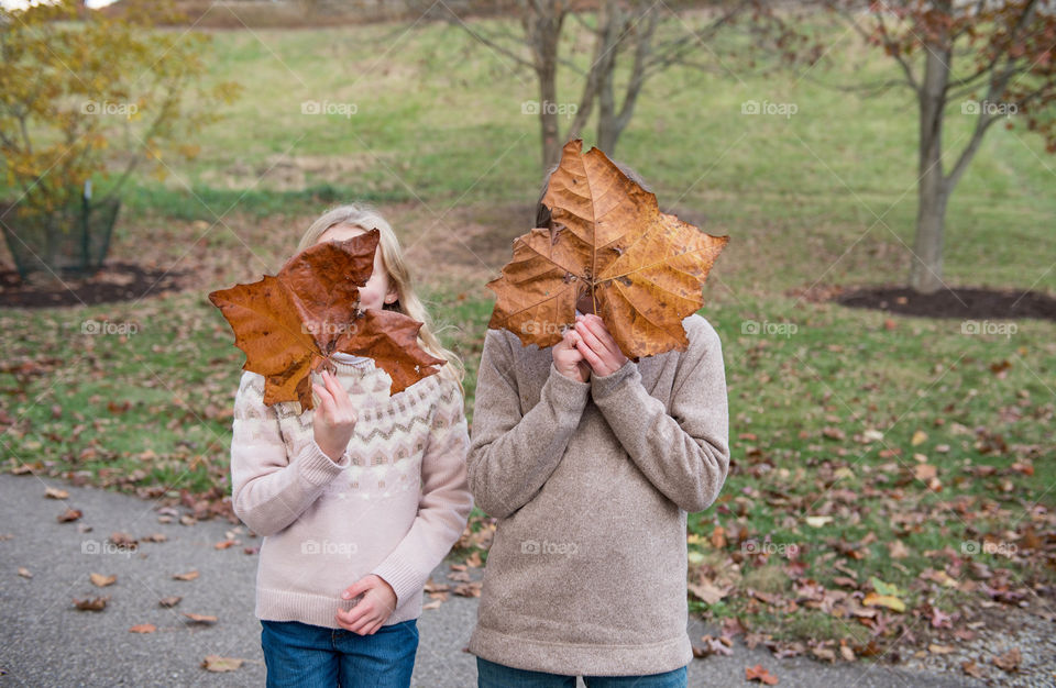 Two children outdoors in autumn covering their faces with large fall leaves 