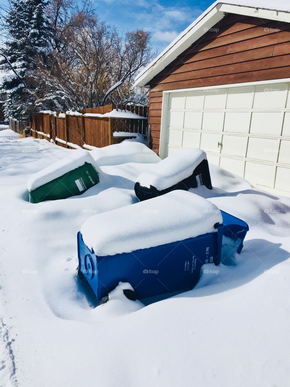 Garbage and recycling containers hidden under the snow outside the house.