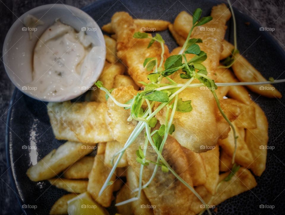 Battered deep fried fish and chips (or fries) freshly prepared on a serving plate as a catering concept. Garnished with sprigs of contrasting greens. A small tub of mayonnaise-based sauce located on one side. A delicious meal.