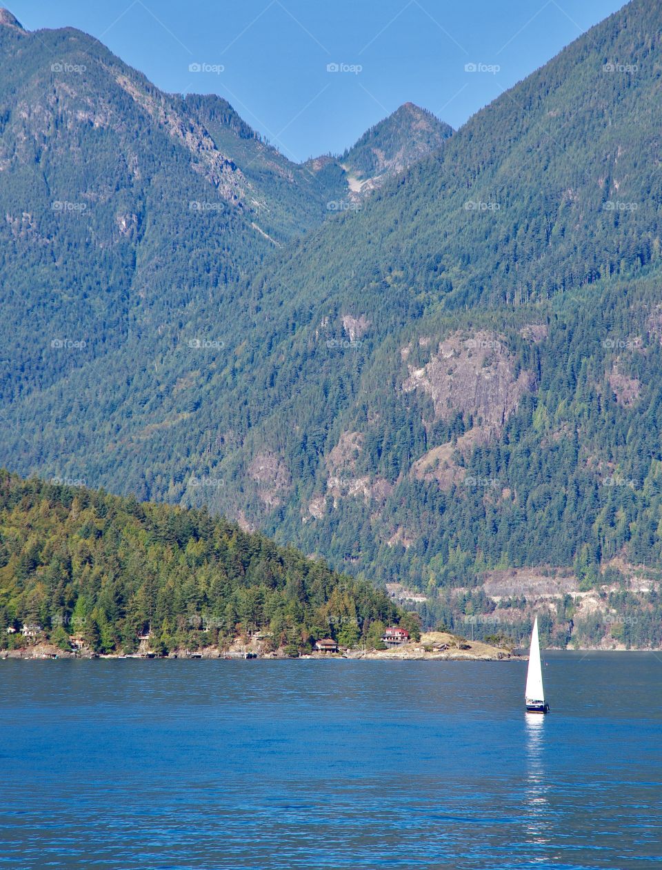 Exterior daylight.  On the ferry near Horshoe Bay, BC, CA.  A single sailboat navigates blue water.  Mountains in background.
