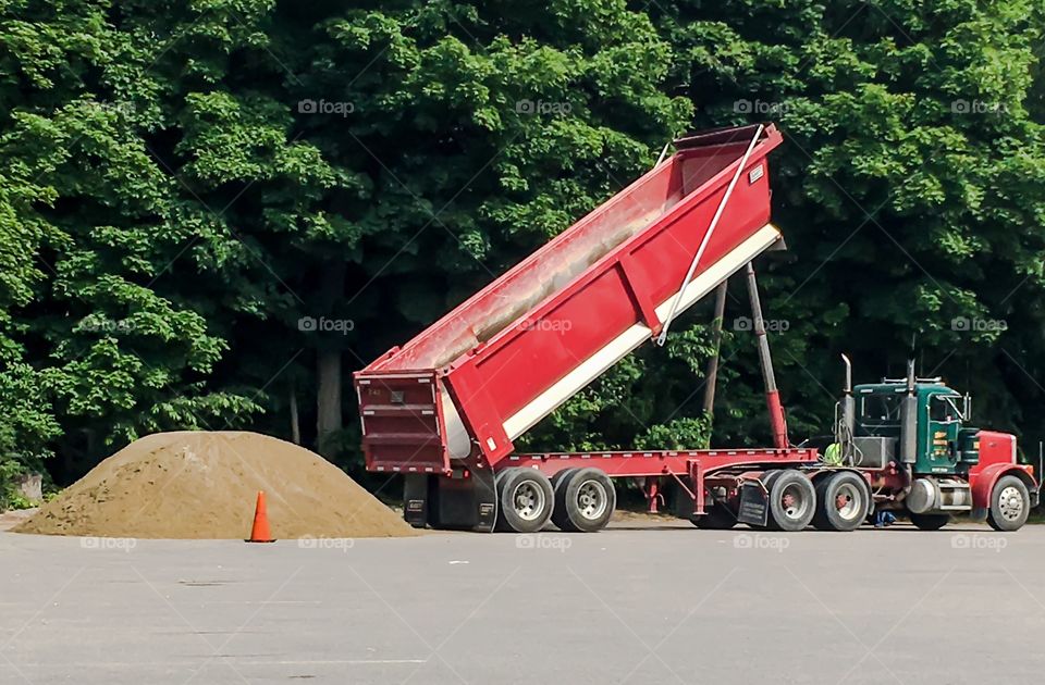 Photo of a truck dumping its load of dirt in a local parking lot.  The truck bed is a vibrant red. 