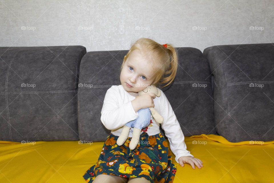 A small, red-haired girl is laughing at home on a gray sofa with purple pillows and a yellow background