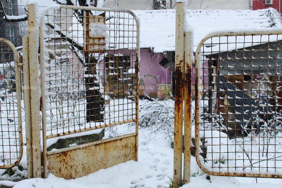 A view of the open old iron fence and the yard of a modest house while the snow is falling