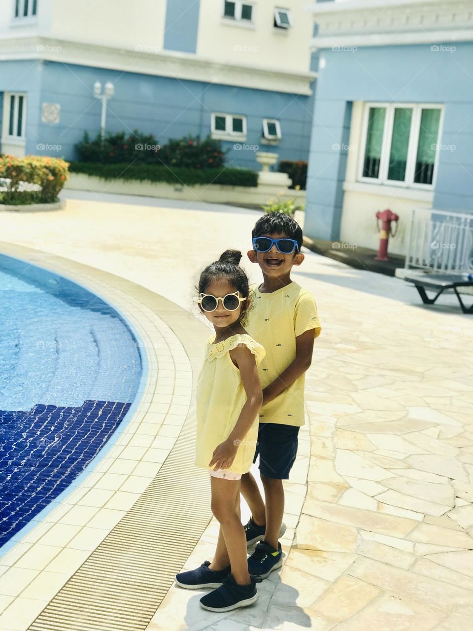 Two kids stand near swimming pool and both wear yellow summer outfits and sunglass.