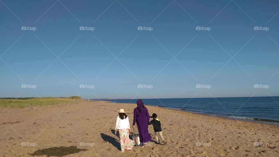 Muslim woman wearing hijab, girl wearing hat and flower skirt and little boy walk on sand beach at Atlantic coast in Cavendish, Prince Edward Island, Canada