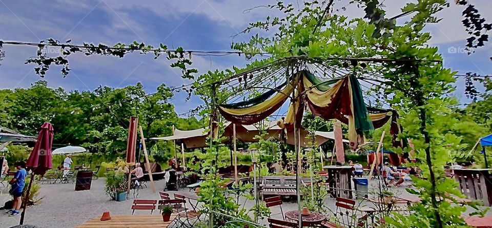 From up closer you can see the wooden deck that accommodates the tables and chairs of the gourmet Asian restaurant café that is located in „München“ or „Munich“ the old „Bavarian“ capitol. 2022. Hypnotic Productions