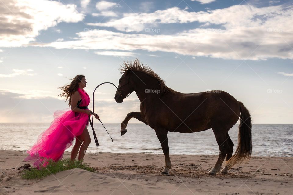 A woman in a pink dress is training horse on the seashore