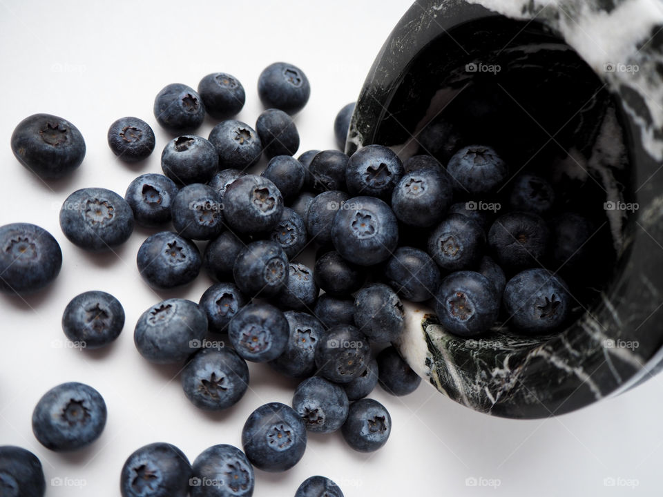 Beautiful blueberries spilling out of a black and white marble bowl over a white table.