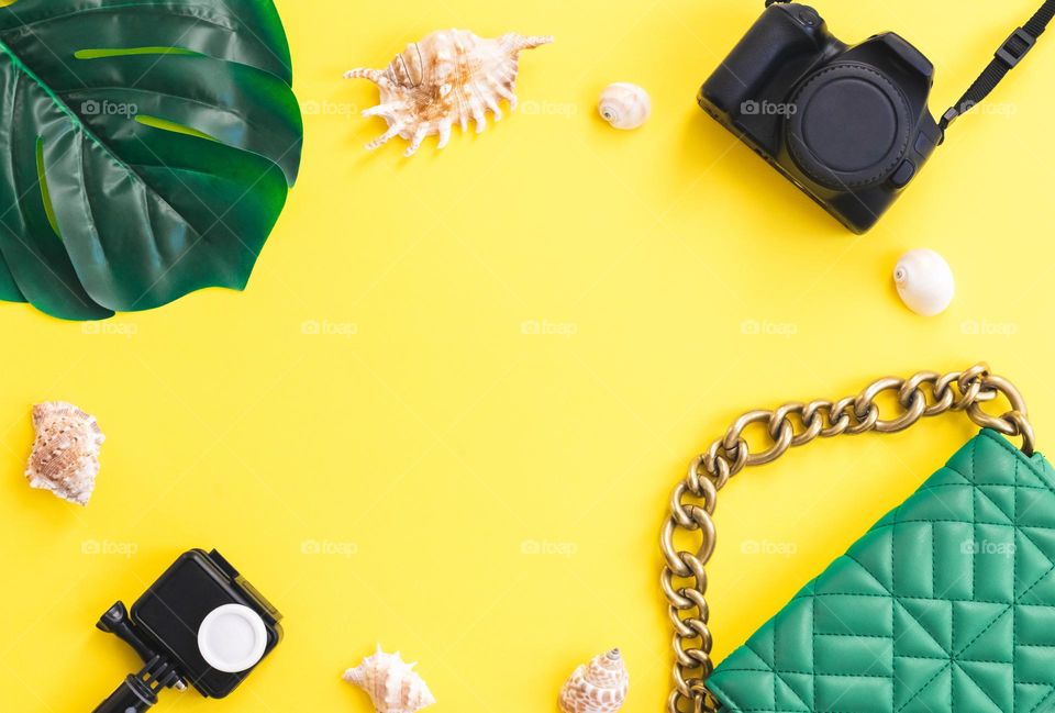 Camera, green fashion quilted bag, action camera, monstera leaf and seashells lie in an oval frame on a yellow background with copy space in the center, flat lay close-up.