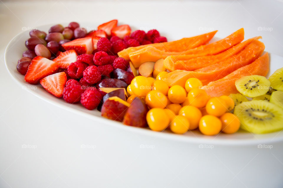 Summer fruit platter with sliced and prepared fresh fruit on a white plate with a white background. Red and yellow fruits include gooseberries, papino, raspberries, strawberries, grapes and plums.