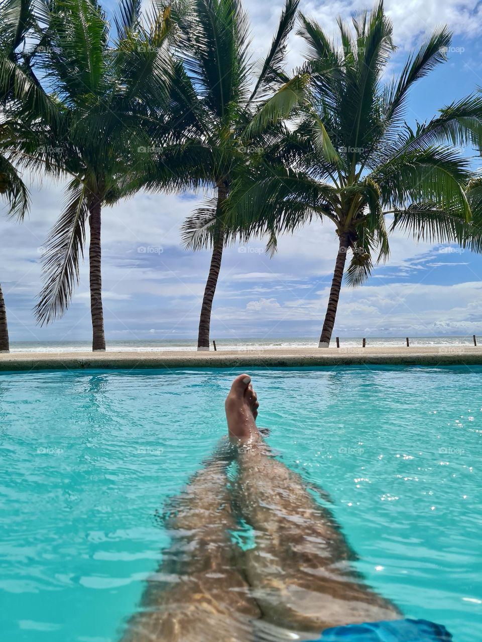 View of the beach and feet, from inside of a swimming pool with palm trees in front of it.