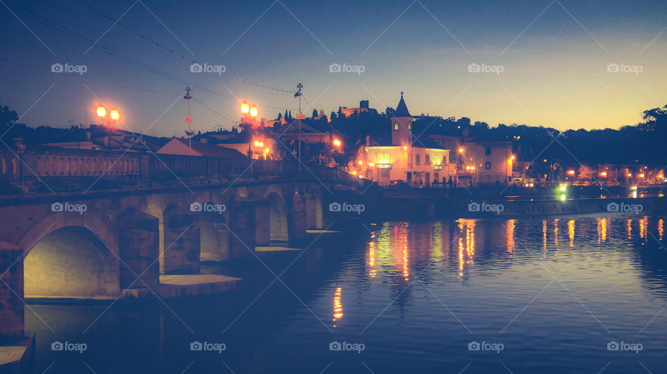 A stone arched bridge over Rio Nabão leads to the lit up view of the old part of Tomar on a clear summer night. The lights of the city reflect in the calm river