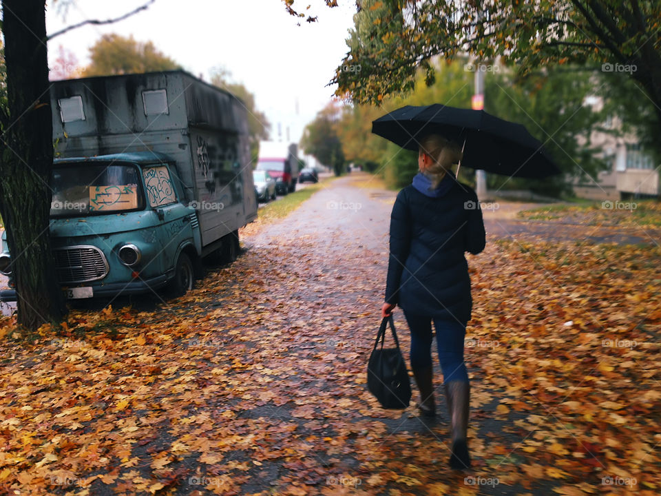 Young woman with headphones walking with umbrella by fallen autumn leaves during the rainy day 
