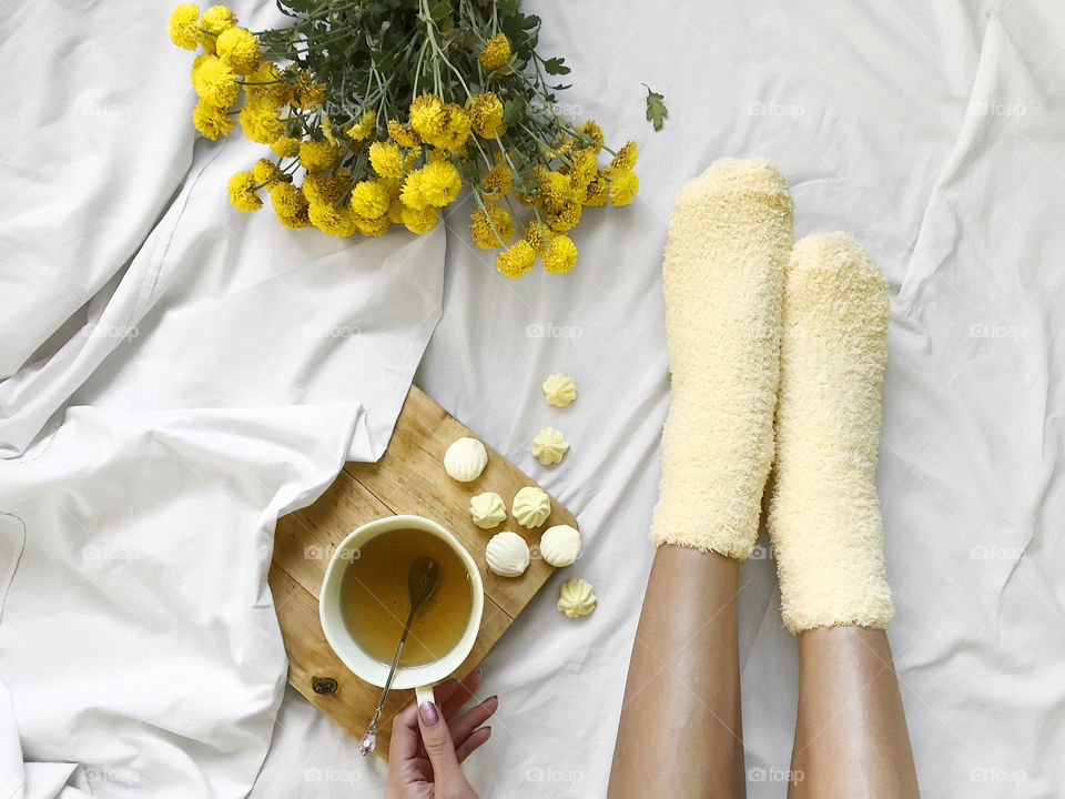 Overheard view of a young woman wearing cozy yellow socks, drinking tea in yellow cup with yellow candies nearby a yellow bouquet of flowers in cozy bed 