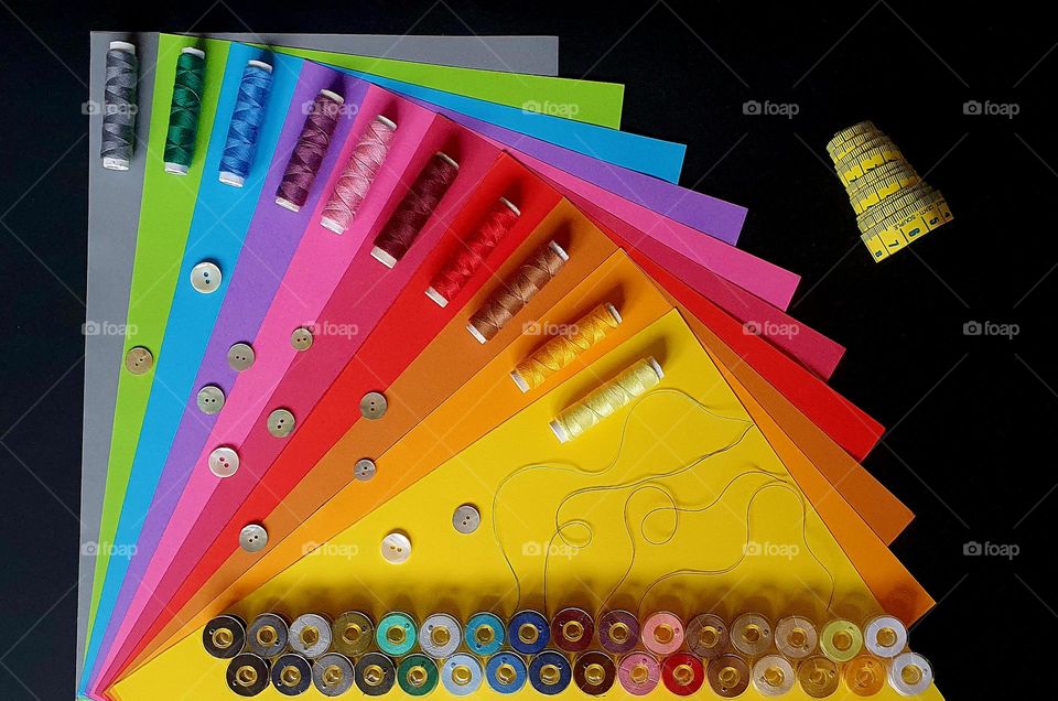 Colourful flatlay on a black surface composed of a fan-shaped set of colour papers on which is placed multi-colour sewing thread rolls and sewing machine bobbins, mother-of-pearl buttons, and yellow measuring tape