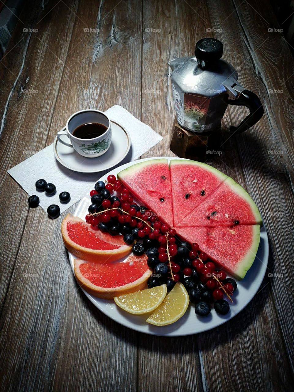 On a wooden background there is a plate with sliced ​​pieces of red watermelon, with bunches of red currants, with black blueberries, with two slices of yellow lemon and with two slices of red grapefruit.  Nearby is a cup of black coffee and a coffee