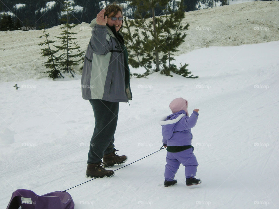 Playtime in the snow! Mom & daughter walking up the tobogganing hill with the sled & waving back at Dad who is waiting at the bottom of the hill.