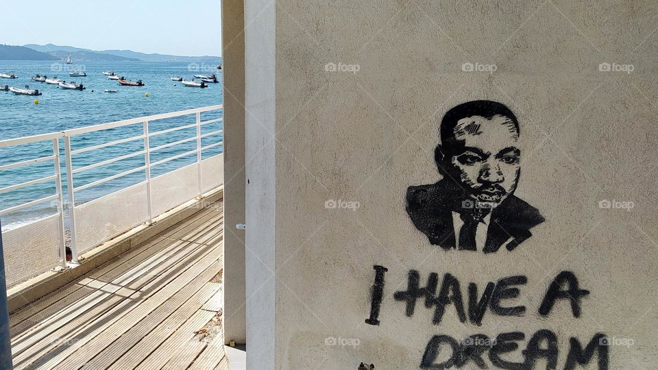 Street art on the way to the beach. This depicts a portrait of Martin Luther King and an inscription of the phrase "I have a dream" from his famous speech. On the left of the photo, the sea and boats at anchor.