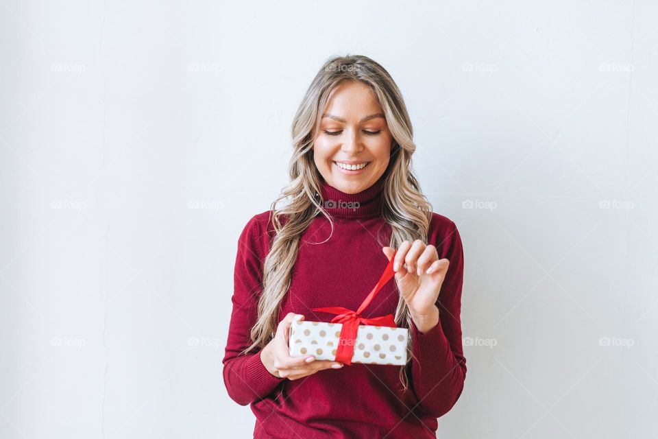 Portrait of happy young adult beautiful woman with blonde long curly hair in bordo sweater with gift box red ribbon in hands on grey background isolated, winter warm clothes
