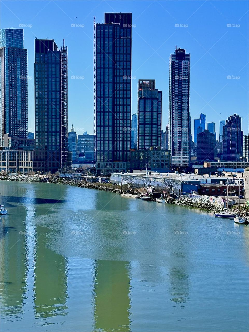 This is „Newtown Creek“ seen from the „Pulaski Bridge“ that connects LIC, Queens to „Greenpoint“, Bklyn. Between two recently erected LIC high rises we see the „NY Life Bldg“ in „Manhattan“ with the golden triangle roof. 2024. Hypnotic Productions