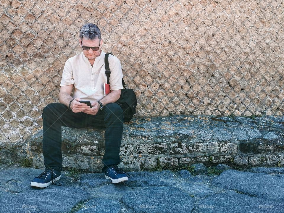 A man with sunglasses sitting beside the wall with camera bag and a map looking on his phone.