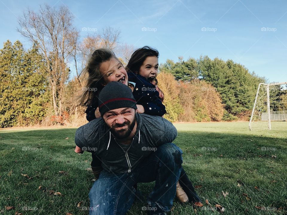 Handsome dad with his two cute little girls on his back playing in the grass at the park with the happiest smiles. 