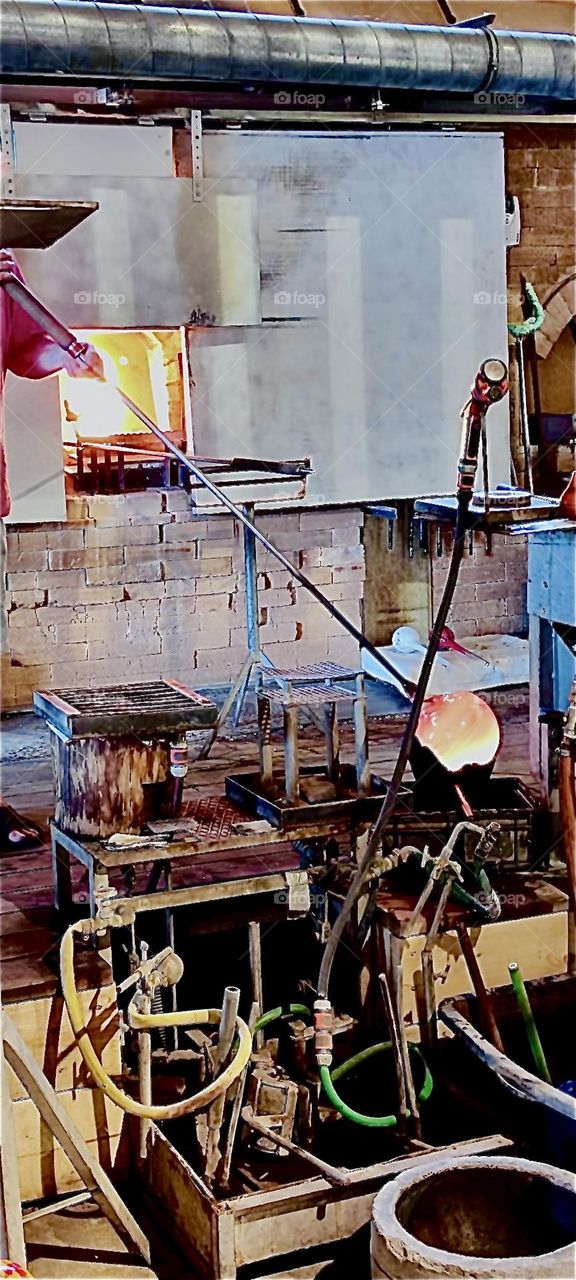 A piece of hot glass at the end of a long thin pipe has been transformed into a hollow sphere by slowly blowing air through it. Where are we ? At a glassblowing workshop in “Freyung”, Bavaria, Germany. 2023. Hypnotic Productions