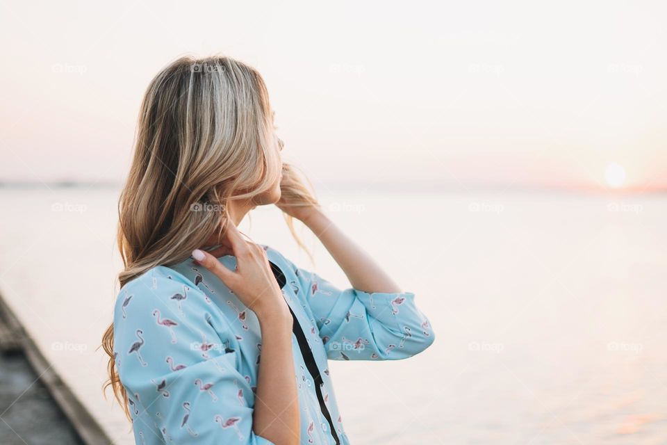 Beautiful blonde young woman in blue dress on the pier and looking on sunset