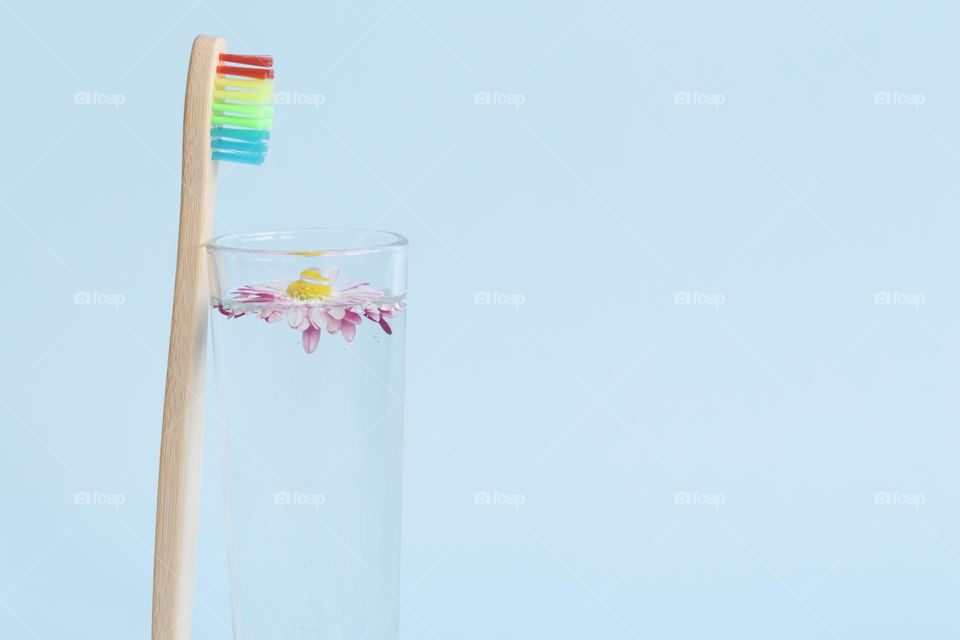 An ecological, wooden and reusable toothbrush with multi-colored bristles stands with a glass of water and a summer daisy, on a blue background.