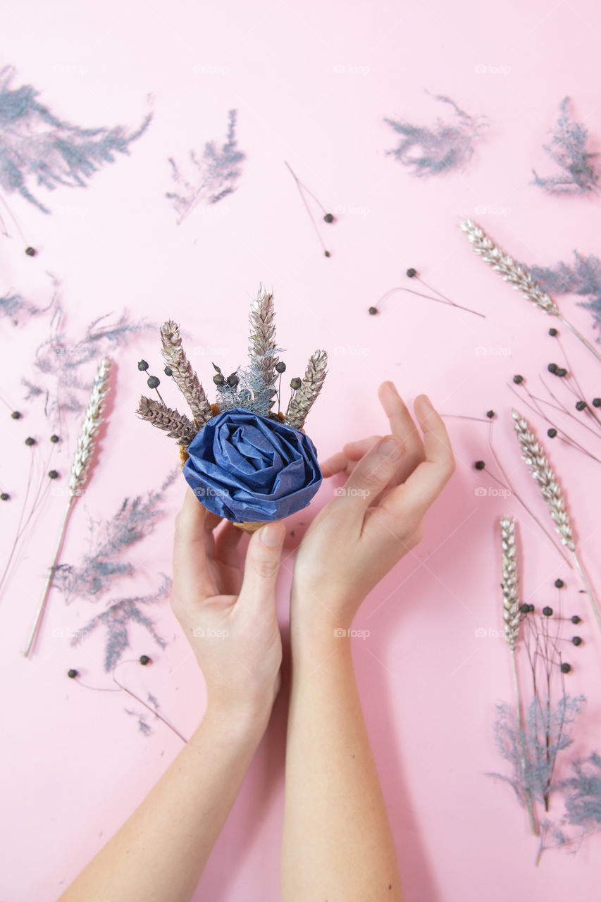 Holiday celebration woman hands holding waffle cone blue handmade rose with dried flowers ,copy paste pink color paper minimalist background.Template greeting card. Flat lay, top view. 